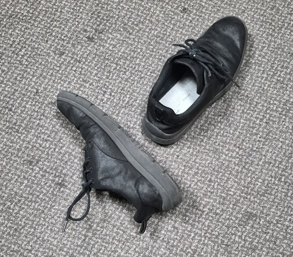 A worn pair of black shoes with a collapsed heel and an uneven sole, the kind that makes the body work harder to stay balanced